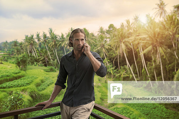 Gutaussehender Mann  der sich auf den Balkon stützt und auf dem Smartphone spricht mit atemberaubender Aussicht auf die tropische Landschaft.