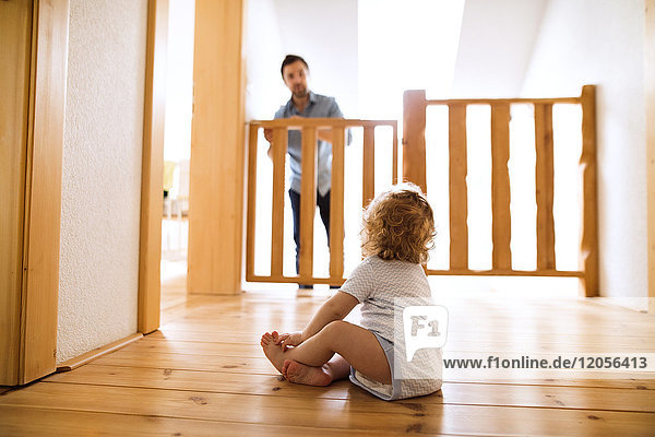 Baby boy sitting on wooden floor looking at father behind barrier at stairs