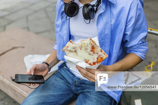 Young man eating pizza on a bench while using cell phone  partial view