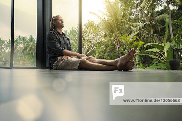 Schöner Mann  der auf dem Boden sitzt und sich an die Glasfassade lehnt  mit einem atemberaubenden tropischen Garten im Hintergrund.