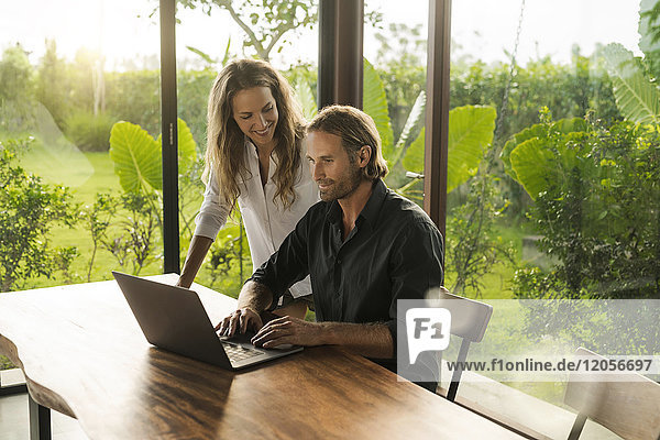 Frau lächelt den Ehemann an  der in einem Designhaus inmitten eines üppigen tropischen Gartens am Laptop arbeitet.
