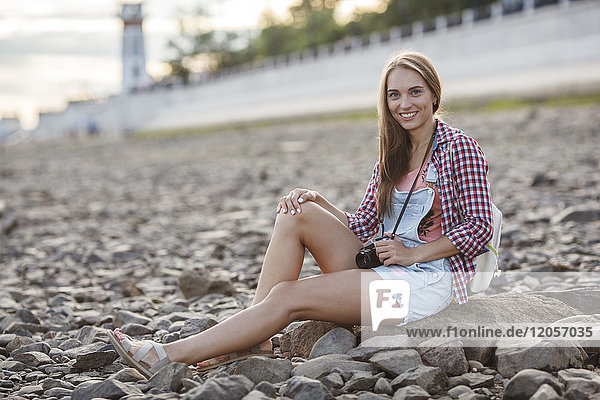 Lächelnde junge Frau mit Kamera am steinigen Strand