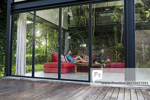 Gartenblick durch Glasfassade des Paares mit Laptop und Buch auf roter Couch im modernen Designhaus