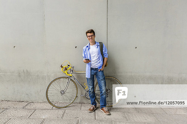 Smiling young man with racing cycle and cell phone in front of concrete wall