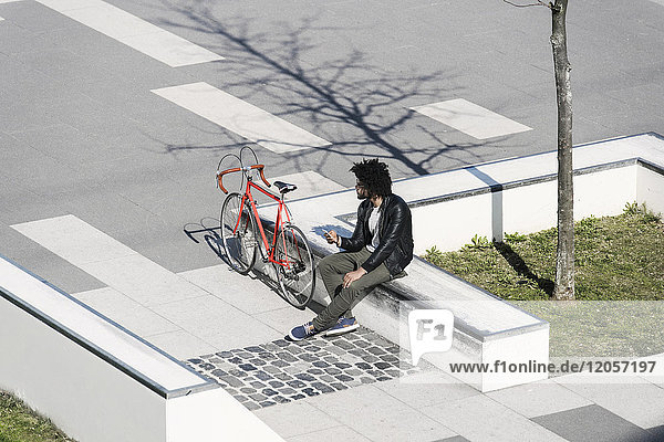Man sitting in city skatepark holding his smartphone next to his bicycle