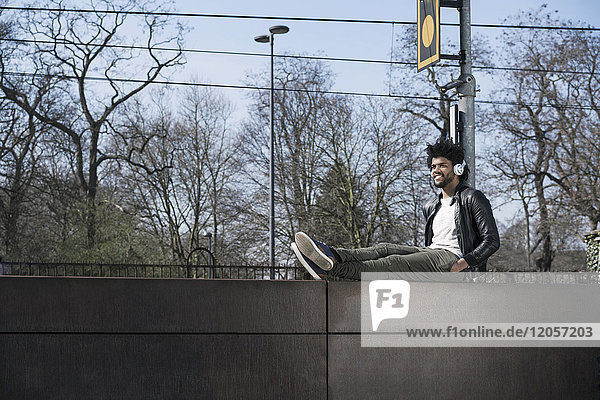 Smiling man sitting on wall listening to music next to train rails
