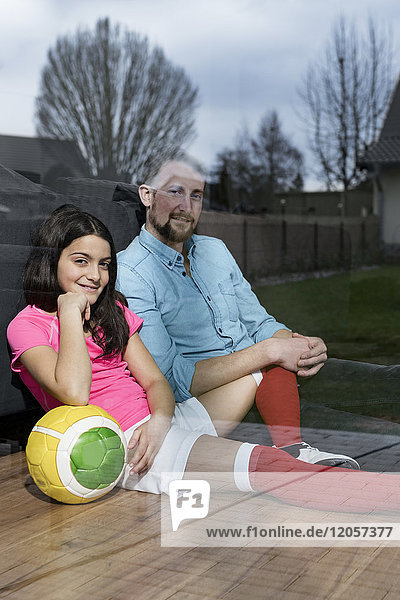 Mädchen im Fußball-Outfit sitzt neben Vater auf dem Boden im Wohnzimmer und schaut aus dem Fenster.