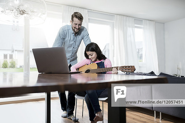Vater lächelt die Tochter an  die vor dem Laptop Gitarre spielt.