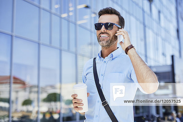 Cheerful businessman talking on phone and holding coffee outdoors