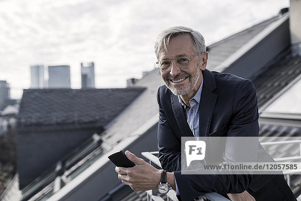 Grauhaariger Geschäftsmann auf dem Balkon hält Smartphone lächelnd in die Kamera