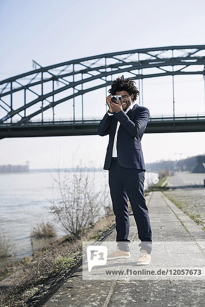 Lächelnder Mann im Anzug am Flussufer beim Fotografieren mit einer Vintage-Kamera
