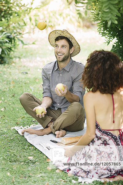 Ein glückliches Paar beim Picknick im Park mit einem Mann,  der mit Äpfeln jongliert.