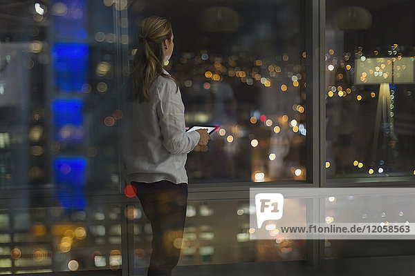 Nachdenkliche Geschäftsfrau mit digitalem Tablet  die spät arbeitet und nachts aus dem Fenster eines städtischen Büros schaut