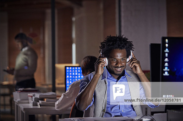Lächelnder Geschäftsmann mit Kopfhörer  der nachts am Computer im dunklen Büro arbeitet.