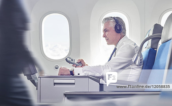 Geschäftsmann mit Kopfhörer und Fernbedienung  der sich einen Film im Flugzeug ansieht.