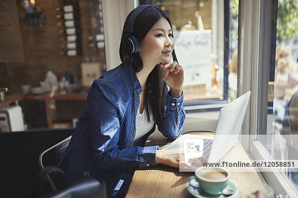 Nachdenkliche junge Frau hört Musik mit Kopfhörern am Laptop und trinkt Kaffee im Caféfenster.