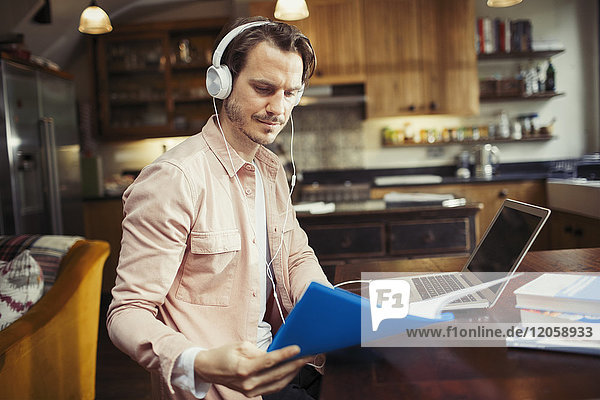 Man with headphones working at laptop  reading paperwork in kitchen