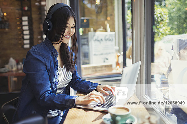 Young woman with headphones using laptop at sunny cafe window