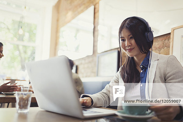 Smiling young woman listening to music with headphones at laptop and drinking coffee in cafe