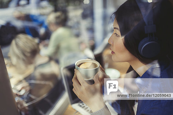 Nachdenkliche junge Frau hört Musik mit Kopfhörern und trinkt Kaffee am Caféfenster.