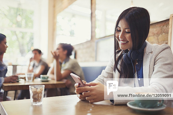 Smiling young woman with headphones texting with cell phone and drinking coffee at cafe table