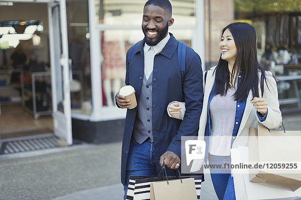 Lächelndes junges Paar geht Arm in Arm entlang der Schaufensterfront mit Kaffee und Einkaufstaschen.