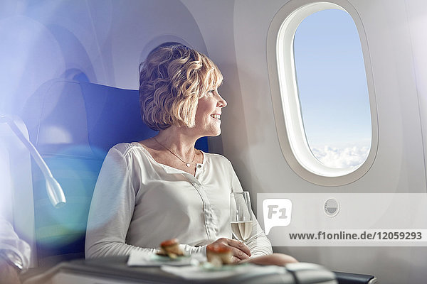 Lächelnde Frau trinkt Champagner  reist in der ersten Klasse  schaut aus dem Flugzeugfenster.