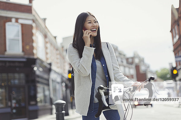 Lächelnde junge Frau pendelt auf dem Fahrrad  telefoniert auf dem Handy in der sonnigen Stadtstraße