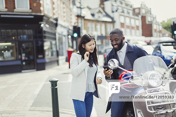 Young couple using cell phone at motor scooter on sunny urban street