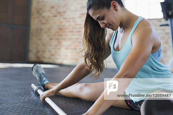 Focused young woman stretching leg  using barbell in gym