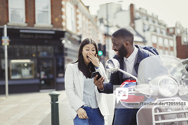 Junges Paar lacht  mit dem Handy am Motorroller auf der sonnigen Stadtstraße
