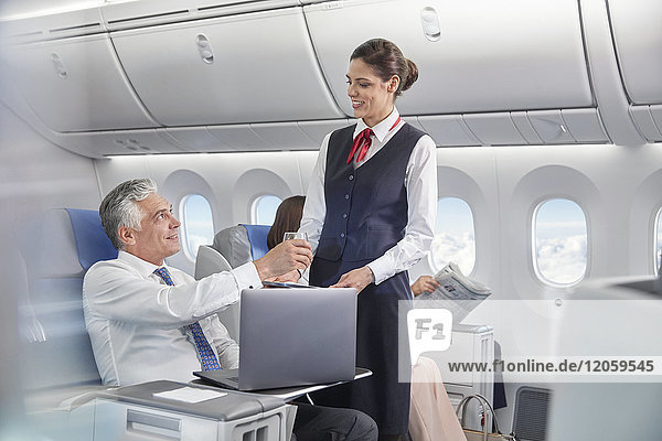 Flugbegleiterin serviert Getränk für Geschäftsmann  der am Laptop im Flugzeug arbeitet