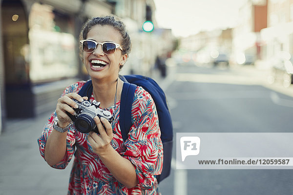 Porträt lachende  begeisterte junge Touristin mit Sonnenbrille fotografiert mit Kamera auf urbaner Straße
