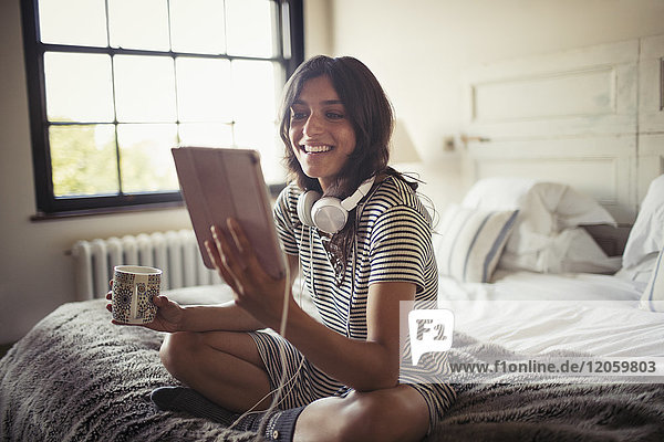 Lächelnde junge Frau mit Kopfhörer  die Kaffee trinkt und ein digitales Tablett auf dem Bett benutzt.