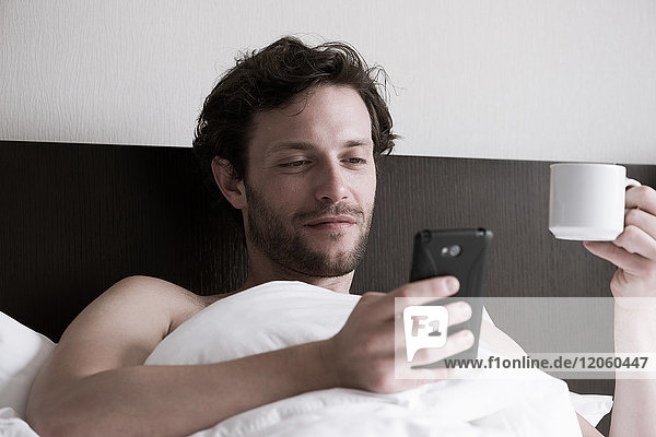Mann im Bett mit Smartphone und heißem Getränk