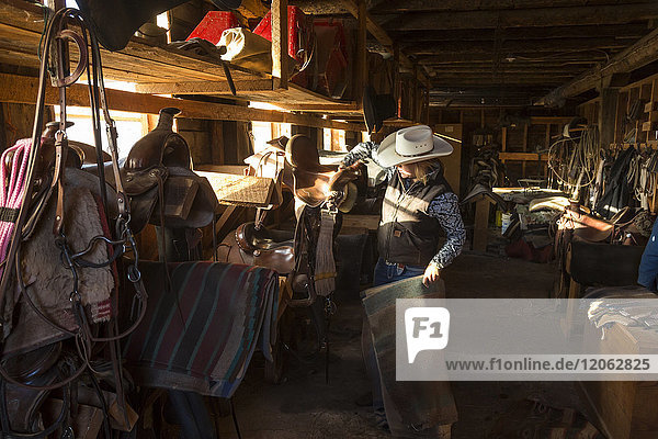 Im Stall stehender Cowboy mit Reitgerten und Sätteln  der einen Ledersattel und eine gestreifte Pferdedecke hält.
