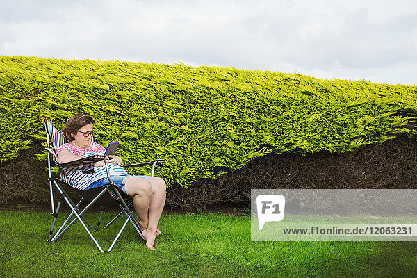 Frau sitzt in einem Campingstuhl an einem geschützten Platz an einer Hecke und benutzt ein digitales Tablett.