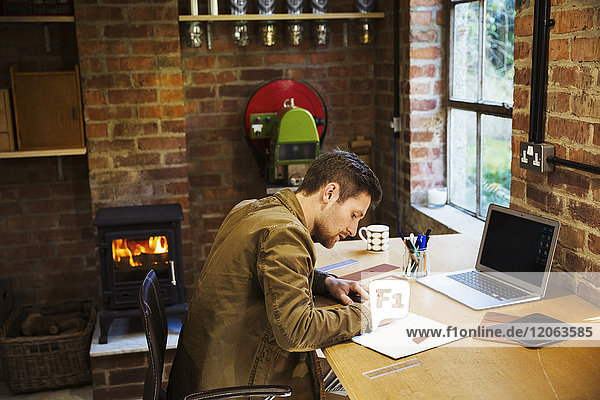 Ein Designer sitzt in seiner Lederwerkstatt an einem Schreibtisch und zeichnet auf Papier. Ein holzbeheizter Ofen mit angezündetem Feuer.