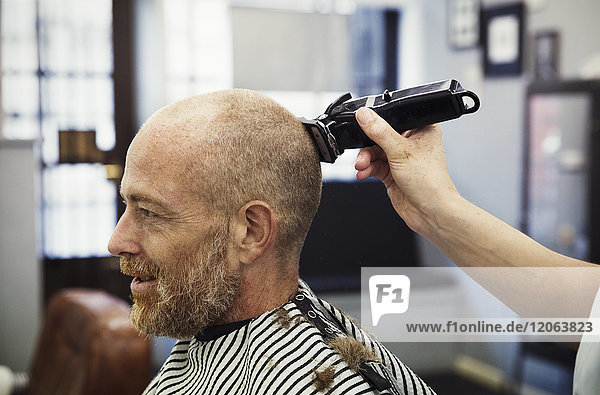 A customer sitting in the barber's chair  and a barber using an electric shaver to shave his head.