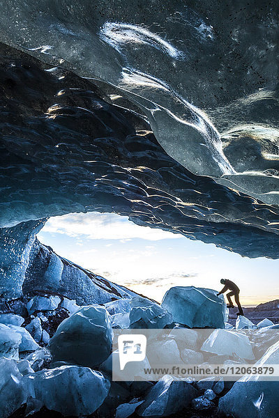 Side view of person climbing up on ice rock at the entrance to a glacial ice cave.