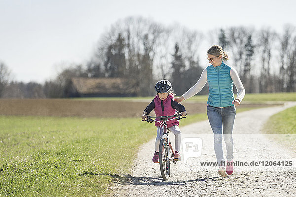 Mother assisting daughter riding bicycle on footpath amidst field