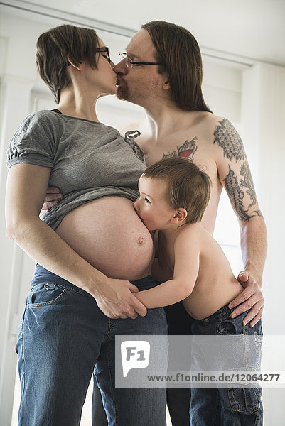 Eltern küssen sich gegenseitig  während der kleine Sohn den Bauch der schwangeren Mutter küsst