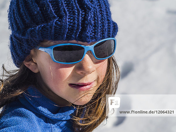 Mädchen mit Sonnenbrille und blauer Strickmütze an einem sonnigen Tag im Schnee