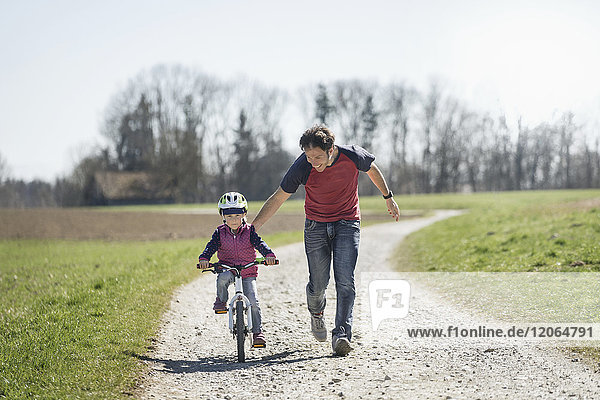 Father assisting daughter riding bicycle on footpath amidst field