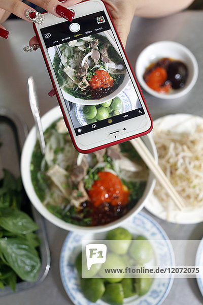 Schüssel mit vietnamesischer Nudelsuppe  bekannt als Pho. Eine Frau fotografiert ihre Mahlzeit mit dem Smartphone. Ho-Chi-Minh-Stadt. Vietnam.
