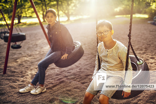 Portrait of brother and sister sitting on swing at park during sunny day