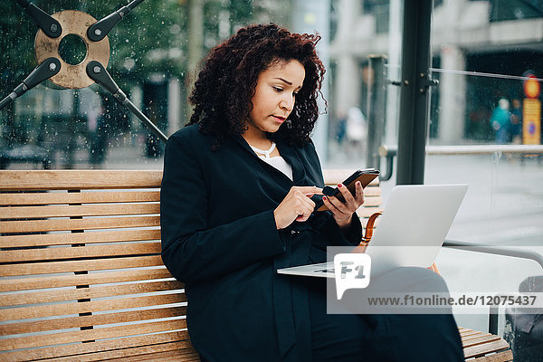 Mid adult businesswoman using mobile phone while sitting with laptop at bus shelter