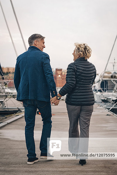 Full length rear view of senior couple holding hands while walking on pier at harbor