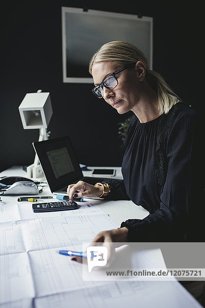 Businesswoman looking at blueprint while using calculator on desk