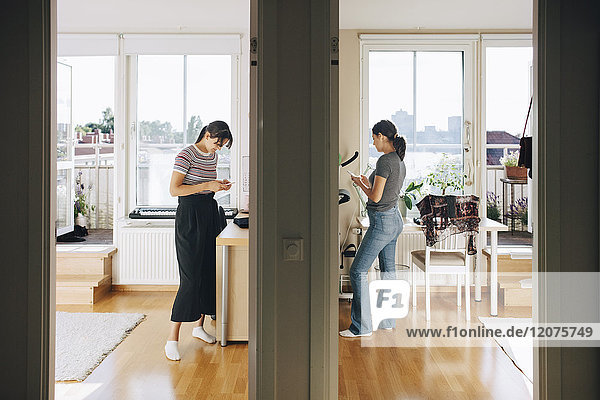 Full length of sisters using mobile phone while standing by wall at home
